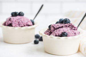 Two small containers of blueberry cheesecake gelato topped with fresh blueberries, each with a spoon.