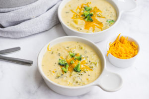 Creamy cheesy broccoli-potato soup on a white serving plate with shredded cheese in another serving bowl.