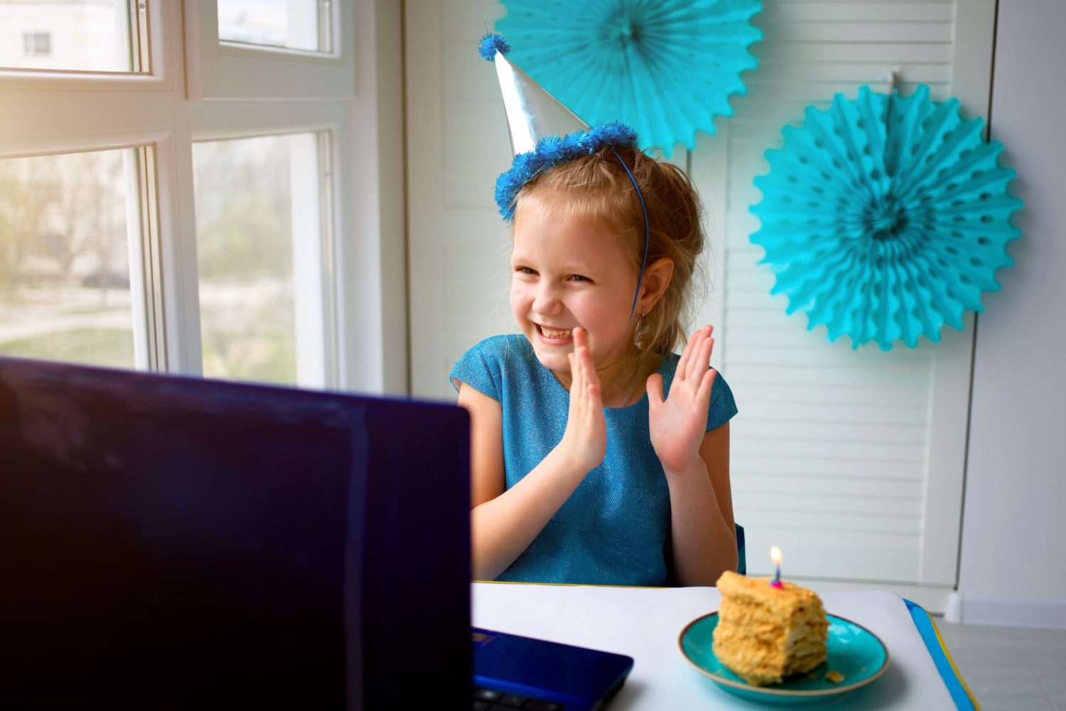 child wearing a party hat and clapping while looking at a computer screen - she has a piece of cake with a birthday candle next to her