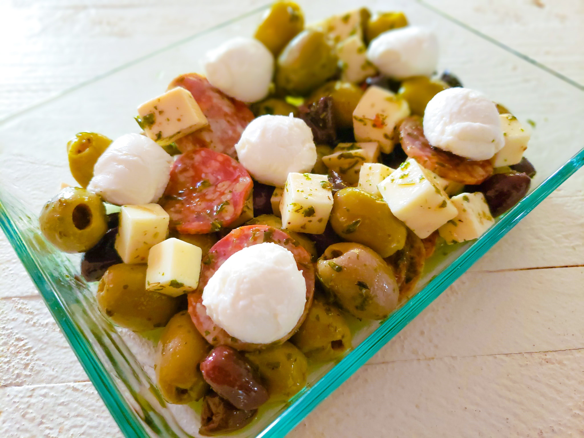 a glass dish with olives, cheese cubes, mozzarella balls and everything has pesto on it