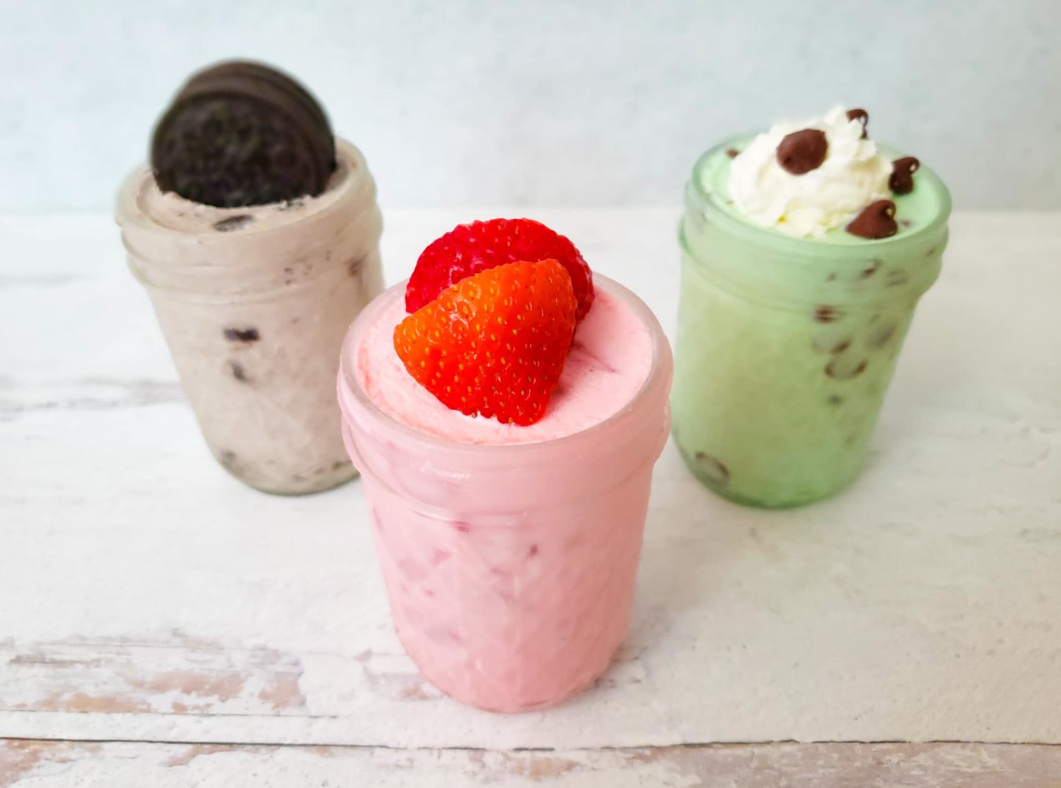 3 small mason jars of ice cream - one with an oreo on top, one with strawberries on top and one with whipped cream and chocolate chips