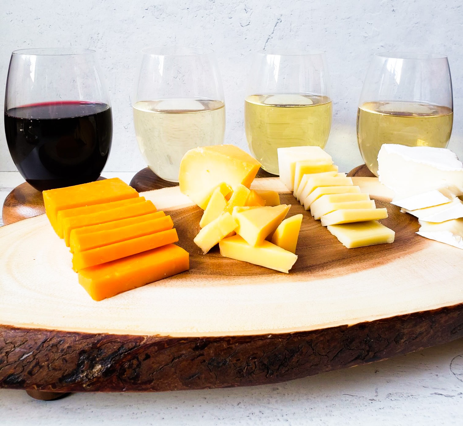 four glasses of wine - 3 white and 1 red are behind a wooden cheese board with 4 kinds of cheese