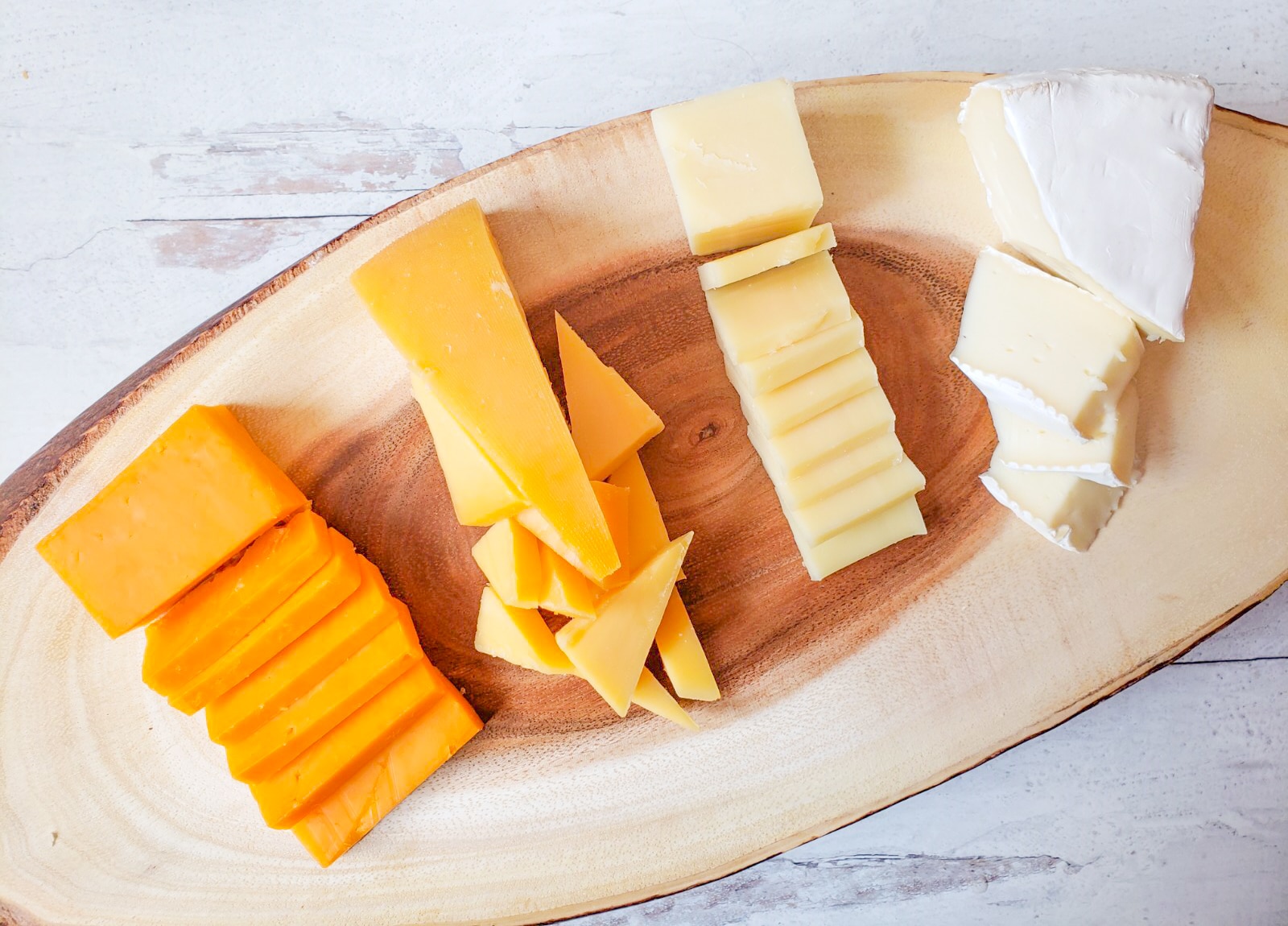 a wooden cheese board with 4 types of cheese sliced and laid on it