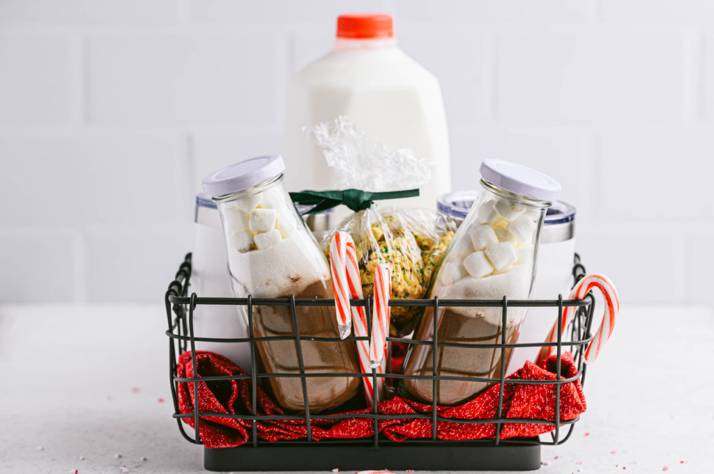 Black basket with two cocoa mugs, marshmallows, candy canes, and a milk jar in the background.