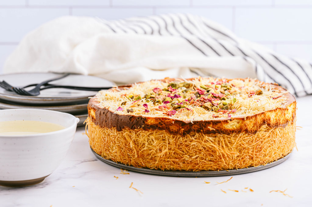 A Knafeh cheesecake decorated with garnishes of ground pistachios, dried rose petals, and rose syrup on a tray.