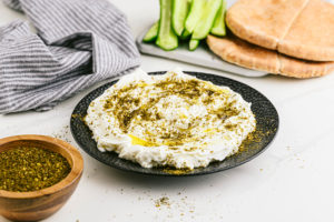 Labna dressed with olive oil and za'atar next to a board with pita bread and cucumbers, a napkin, and za'atar.