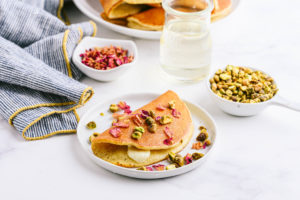 Qatayef on a plate garnished with pistachio and rose petals ladled with syrup next to a napkin.