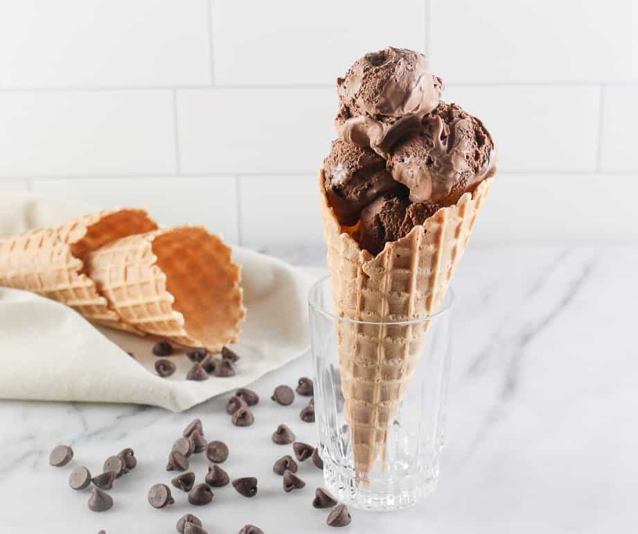 Un cono con helado de cuatro chocolates colocado sobre un vaso de cristal, junto a un par de conos sobre una servilleta.