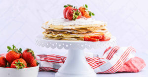 Una tarta de crepa de creama de fresas servida sobre una base para pastel blanca, decorada con fresas.