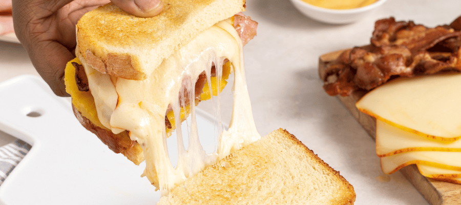 A hand holds a slice of a grilled cheese sandwich with melty cheese stretching from one slice to the other.