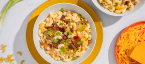 A bowl of mac and cheese with bacon and jalapeño on a yellow plate next to a plate with cheddar cheese.