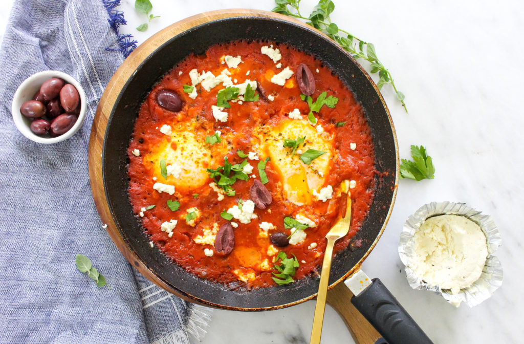 A skillet brimming with delectable greek shakshuka eggs, complemented by olives and feta cheese in the background.