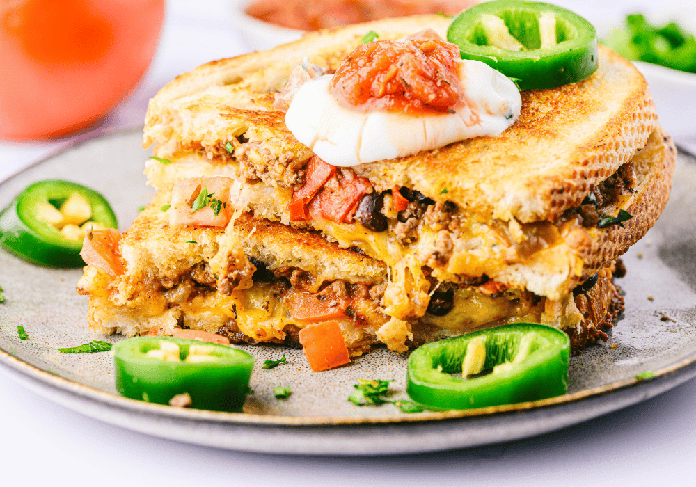 A plate with nacho grilled cheese cut in half decorated with pepper jack slices, salsa, and sour cream.
