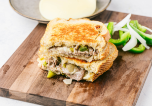 A cut half philly cheesesteak grilled cheese on a wooden table, with chopped onions and jalapenos in the background.