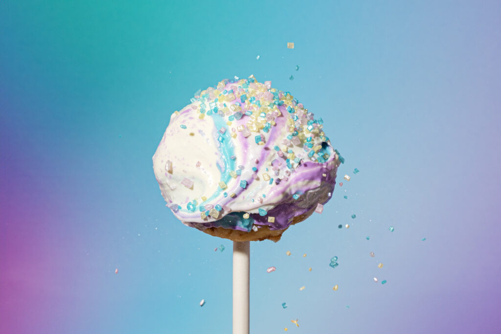 A singular galaxy cake pop close up with colorful icing and sprinkles falling off.