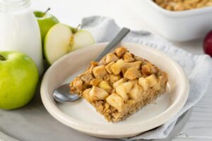 Un trozo de tarta de manzana servido en un plato junto a un plato y manzanas verdes Granny Smith.