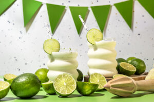 Two tropical lime cocktails in curved glasses with lime wheels and striped straws, surrounded by whole and halved limes on a bright green surface.