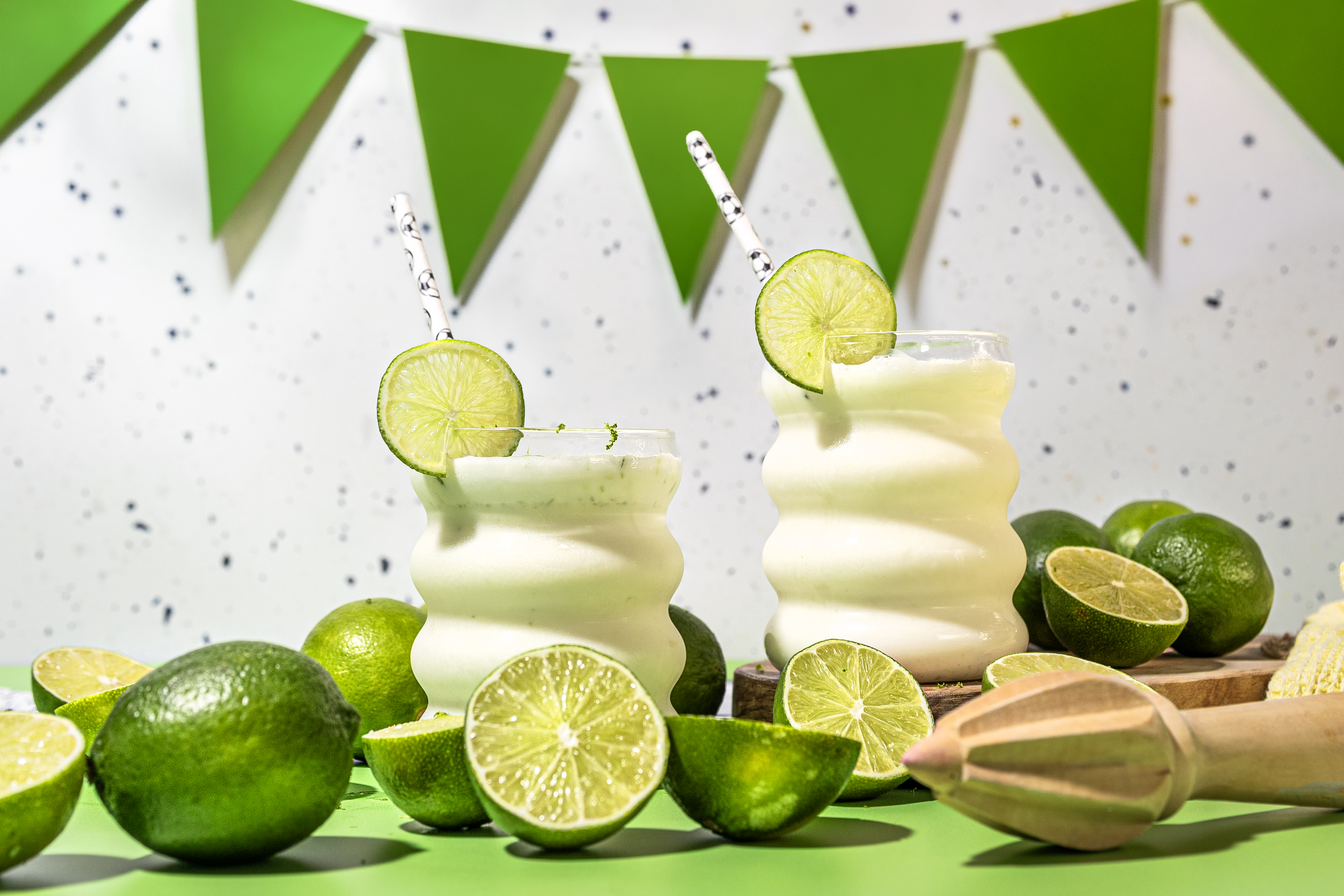 Two tropical lime cocktails in curved glasses with lime wheels and striped straws, surrounded by whole and halved limes on a bright green surface.