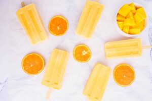 Mango-Orange-Yourt-Popsicles_10-rotated-1 | Savor Recipes Assorted orange popsicles arranged with orange halves and a bowl of mango chunks on a white parchment background.
