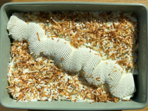 Rectangular baking dish with white frosting, toasted coconut flakes, and ribbed meringue rosettes while resting on a wooden surface.