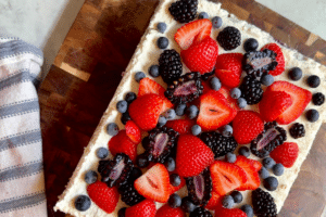 Cream-topped fruit cake slice topped with fresh strawberries, blueberries, and blackberries on a wooden board beside a striped cloth.