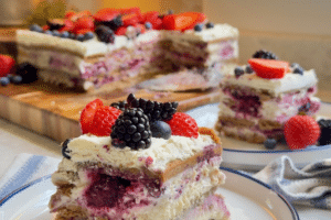 Layered berry cake slice on a white plate, topped with strawberries, blueberries, raspberries, and blackberries.